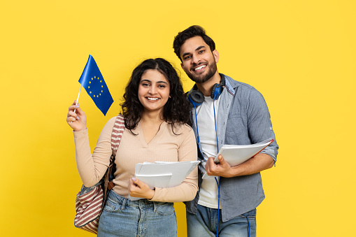 Happy Young Students Holding European Union Flag With School Supplies Over Yellow Background
