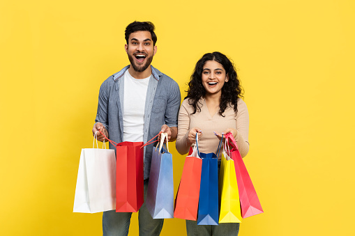 A Joyful Young Couple Smiling As They Hold A Variety Of Colorful Shopping Bags Against A Bright Yellow Background, Reflecting The Excitement Of Retail Consumerism And Fashionable Purchases.