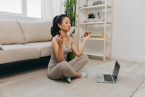 Asian woman in tracksuit sits with laptop in lotus position at home and meditates with eyes closed with a slight smile, mental health support lifestyle. High quality photo