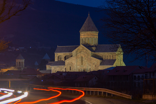 Night view of the Svetitskhoveli Cathedral, Mtskheta, Georgia. Illuminated with warm light against a dark blue night background. A part of the road with white and red traces of car lights is visible.