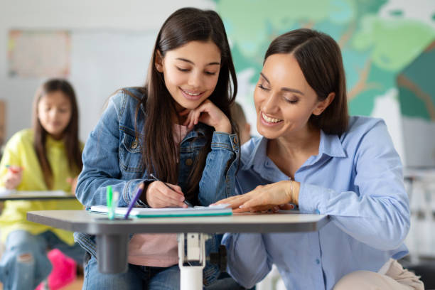 Young teacher explaining something to preteen girl, studying, learning language with school children sitting in classroom