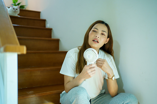 Unhappy Asian woman using a portable electric fan blowing herself. Sweaty Asian woman relaxing at home during the hot weather day and using portable electric hand fan.