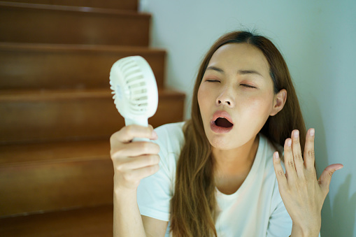 Unhappy Asian woman using a portable electric fan blowing herself. Sweaty Asian woman relaxing at home during the hot weather day and using portable electric hand fan.