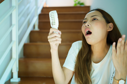 Unhappy Asian woman using a portable electric fan blowing herself. Sweaty Asian woman relaxing at home during the hot weather day and using portable electric hand fan.