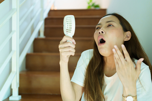 Unhappy Asian woman using a portable electric fan blowing herself. Sweaty Asian woman relaxing at home during the hot weather day and using portable electric hand fan.