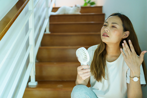 Unhappy Asian woman using a portable electric fan blowing herself. Sweaty Asian woman relaxing at home during the hot weather day and using portable electric hand fan.