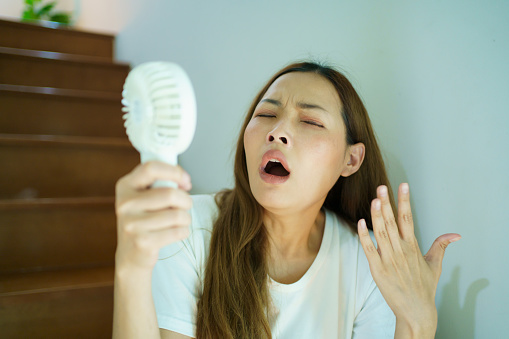 Unhappy Asian woman using a portable electric fan blowing herself. Sweaty Asian woman relaxing at home during the hot weather day and using portable electric hand fan.