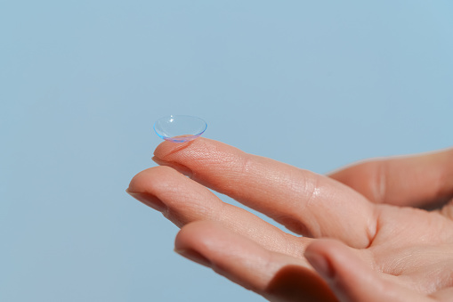 Finger of female hand holding transparent contact lens on blue isolated background. Concept of ophthalmology, improvement of vision