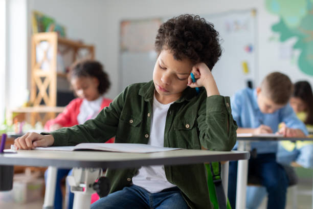 istock Tired and bored boy sitting at desk in classroom at school, writing in notebook and thinking, resting head on hand 2148801242
