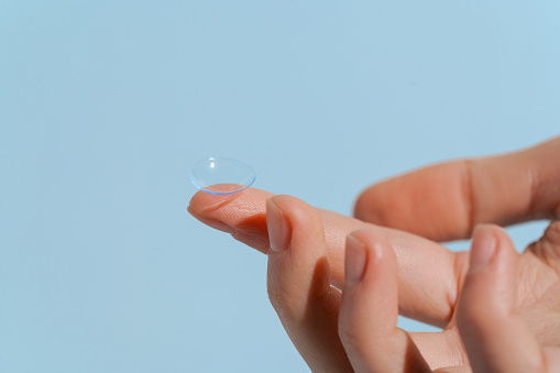 A finger of a female hand holds a blue transparent contact lens on a blue isolated background. Vision improvement concept, ophthalmology, farsightedness
