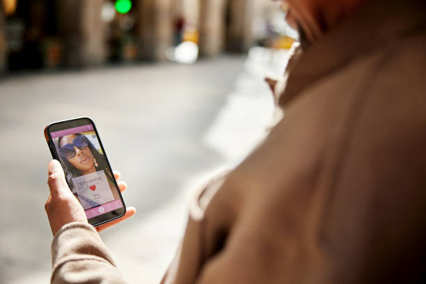 Smiling man looking at dating app message, it’s a match Over the shoulder view with focus on smart phone with profile picture of mature woman wearing sunglasses. heart emoji phone stock pictures, royalty-free photos & images