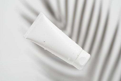 White facial cream tube mockup with water drops on palm branch shadow background on gray isolated background. The concept of moisturizing and nourishing the skin.