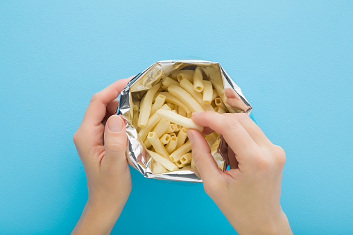 Young adult woman hand taking white sticks of potato chips from opened foil bag on light blue table background. Pastel color. Closeup. Point of view shot. Top down view.