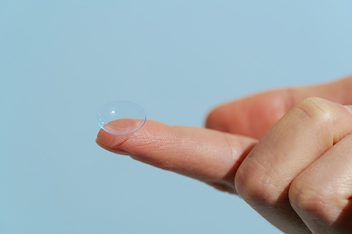 Finger of female hand holding blue transparent contact lens while wearing on blue isolated background. Vision improvement concept, farsightedness and nearsightedness, ophthalmologist.