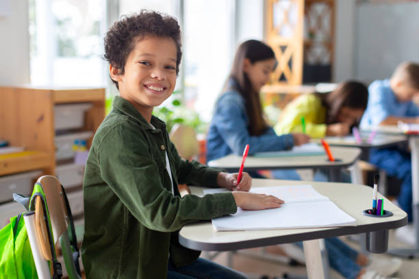 istock Smiling black boy writing in classroom, learning, knowledge and study. Latin male pupil at school, preparing for exam test and project 2148802179