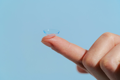 A finger of a female hand holds a blue transparent contact lens on a blue isolated background. Concept of ophthalmology, myopia, farsightedness, treatment of disorders, medicine.