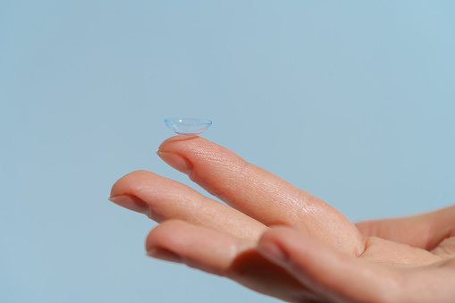 female hand holding blue clear contact lens before putting on eye on blue isolated background. Vision improvement concept, myopia.