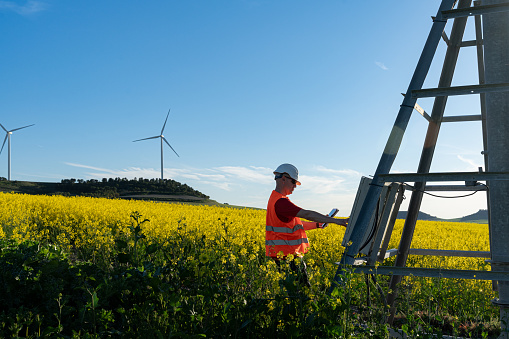 In a canola field in full bloom an agricultural irrigation technician is repairing a sprinkler irrigation tower, the technician manipulates the control panel of the irrigation station while consulting data on the digital tablet he is holding in another hand. In the background two wind turbines on a small hill.