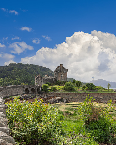 Dornie - United Kingdom. May 22, 2023: Vertical view of Eilean Donan Castle, a Scottish stronghold, under a canopy of billowing clouds, the Scottish flag whispering above its battlements