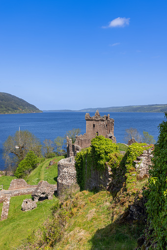 Drumnadrochit - United Kingdom. May 25, 2023: A vertical perspective showcases Urquhart Castle ruins overlooking the tranquil Loch Ness in Scotland, with verdant foliage enveloping ancient stone walls