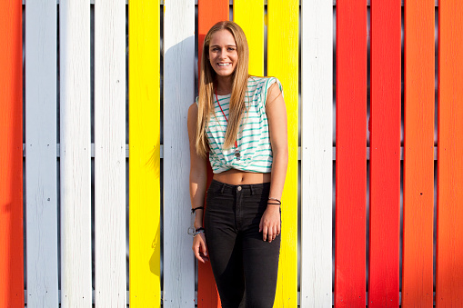 Stylish beautiful young woman poses in front of a vibrant colorful wooden wall on street, outdoors during summer on the beach. Urban style.