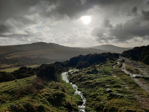 Cloudy skies with sun shining through over Dartmoor. Hills and Tors in the distance and river running alongside footpath. Lots of very green open moorland