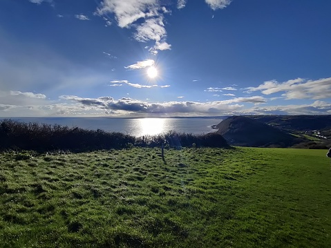 Sunny day with blue skies reflected in blue sea. Taken from top of cliffs on footpath between Beer and Branscombe with green grass heading down the hill. Lovely reflection of sun in the sea