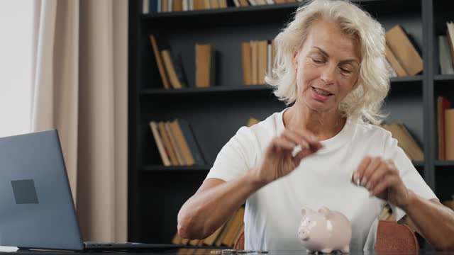 Senior woman puts coins in piggy bank. Happy smiling old lady gray haired female putting coins in pink piggy bank at home. Budget planning, expenses. Saving money, self finance, deposit concept.