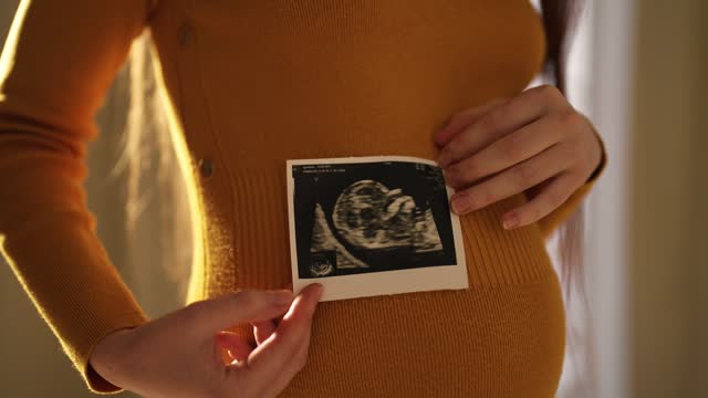 Pregnant woman looking ultrasound report. Happy pregnant female watching her ultrasound report and touching her abdomen, admiring sonography picture of her baby. Expecting baby, pregnancy concept.