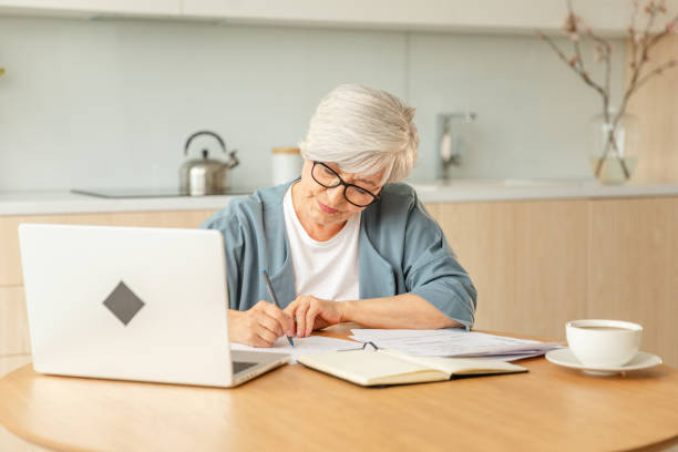 istock Middle aged senior woman using laptop computer writing notes at home. Focused mature old businesswoman work in home office making written records doing online work. Mature boss professional worker. 2148813898
