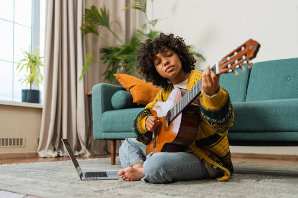 istock African American woman learning guitar watching tutorial on laptop at home. Black girl playing guitar singing song learning online music lessons, Artistic woman playing acoustic guitar learning chords 2148814005