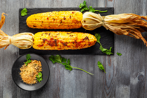 The cob fried corn on a white plate with sliced grilled onions and herbs on white wooden table.