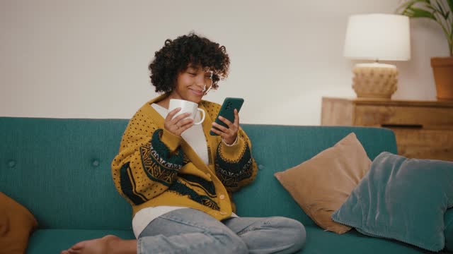 Young girl with phone drink tea. African American beautiful woman hold in hand use mobile phone drink coffee. Girl relax spend free spare time in living room with phone. Communication with friends.