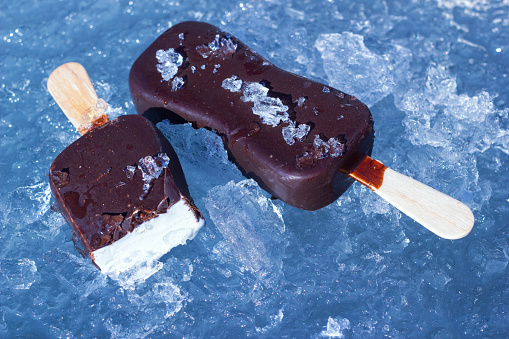 The classic milky ice cream with chocolate glaze on wooden stick. The sweet frozen dessert on the natural chopped ice. Closeup view.