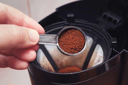 The process of filter coffee preparation. Portion of ground coffee spilled in electrical dripper. Closeup view with selective focus on spoon.
