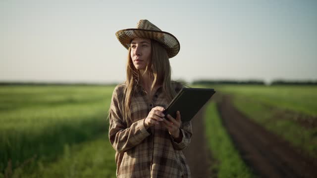 Farm worker woman with tablet in hands walks on agricultural field early spring at sunset. Blonde caucasian female works on farmland outdoors. Food production, farming country, agribusiness concept.