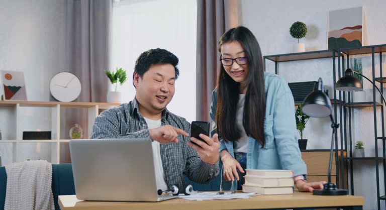 Smiling asian couple man and woman talking and using smartphone while working together on laptop over startup project