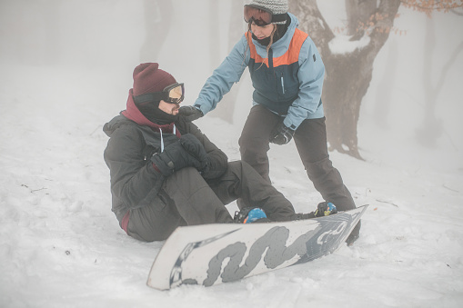 A male snowboarder sits on the snow after a fall, while another snowboarder approaches him and offers help