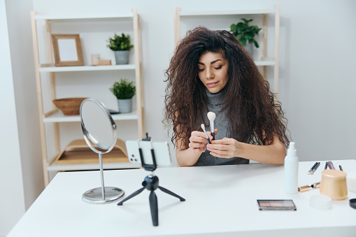 Makeup school concept. Professional visage artist tanned handsome curly Latin beauty blogger in casual top in home interior. Copy space Banner. Influencer record video review blog using smartphone