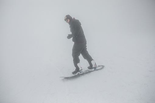 A snowboarder with gear is snowboarding on a mountain during a foggy day. He performs acrobatics and enjoys the adrenaline that this extreme winter sport provides