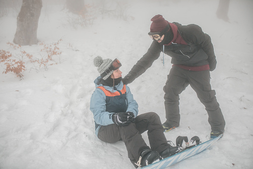 A female snowboarder sits on the snow after a fall, while another snowboarder approaches her and offers to help her