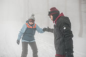 A man teaches a woman to snowboard on a foggy, winter day. He holds her hand and helps her with snow activities