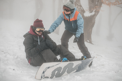 A male snowboarder sits on the snow after a fall, while another snowboarder approaches him and offers help