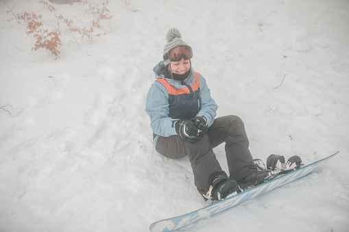 A woman sits on the snow after falling from a snowboard. She has a smile on her face because the fall wasn't serious, it was more adrenaline and fun