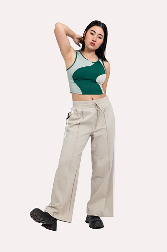 A full-shot portrait of a young woman standing in front of a creme background in a studio in Newcastle upon Tyne. She is touching her hair. She is wearing a crop top and beige trousers. Videos are available similar to this scenario.