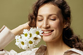 Close up portrait of smiling curly female in linen casual shirt hold chamomiles flowers near face close eyes posing isolated on over olive green pastel background. Nature is beauty concept. Copy space