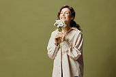 Smiling cute curly beautiful female in linen casual shirt hold chamomiles flowers near face enjoying smell posing isolated on over olive green pastel background. Nature is beauty concept. Copy space