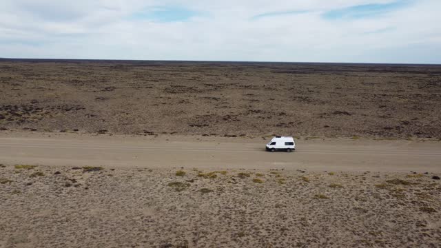 Aerial video side view of small white van driving on gravel road on flat arid landscape