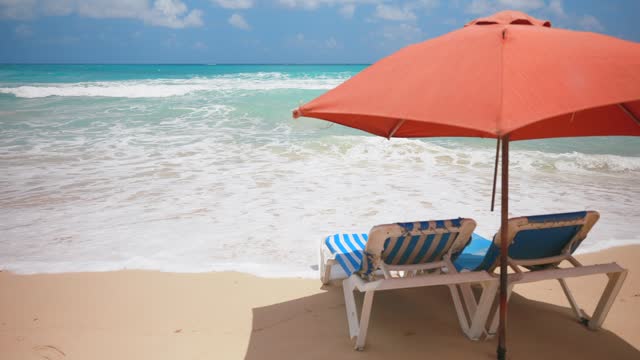 Slow motion of two empty blue sun loungers under the red parasol on the sandy beach near the sea water with big waves. Rest and relaxation summer holiday scene