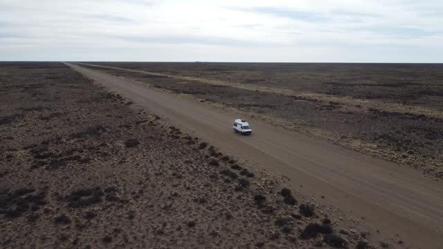 Aerial video circling a small white van driving on gravel road on flat arid landscape